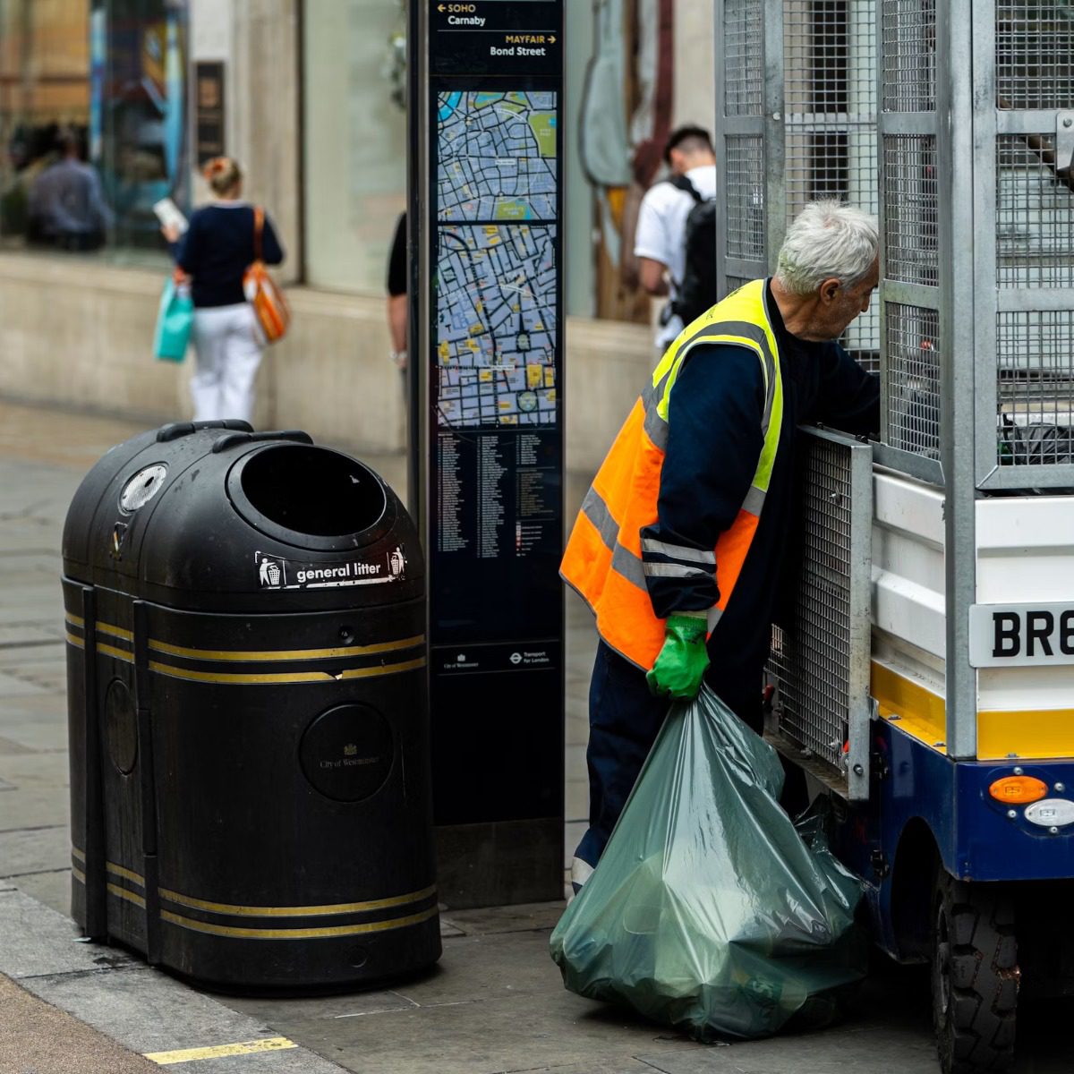 Municipal sanitation worker loading collected street litter into a maintenance vehicle beside a public trash bin, representing municipal street sweeper services near me.